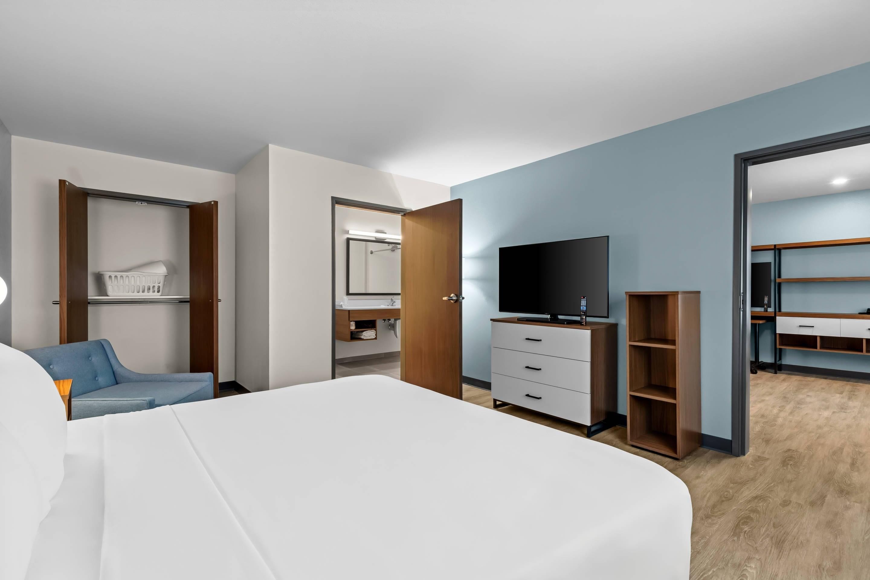 A modern hotel room with a large bed, a television, a wardrobe, and a bathroom visible through an open door.