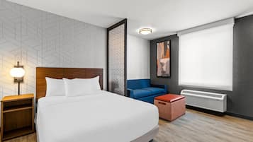 A modern hotel room with a large bed, a nightstand, a blue sofa, a red ottoman, a wall-mounted heater, and a geometric-patterned wall.