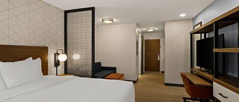 A modern hotel room with a large bed, a desk with a computer, a TV, and a seating area with a chair and a small table.