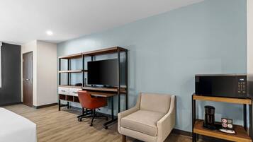 A hotel room with a bed, a desk with a computer and TV, a chair, a coffee maker, and a microwave on a shelf.