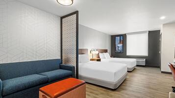 A hotel room with a sofa, two beds, a small table, a wall with geometric patterns, a ceiling light, and a window with blinds.