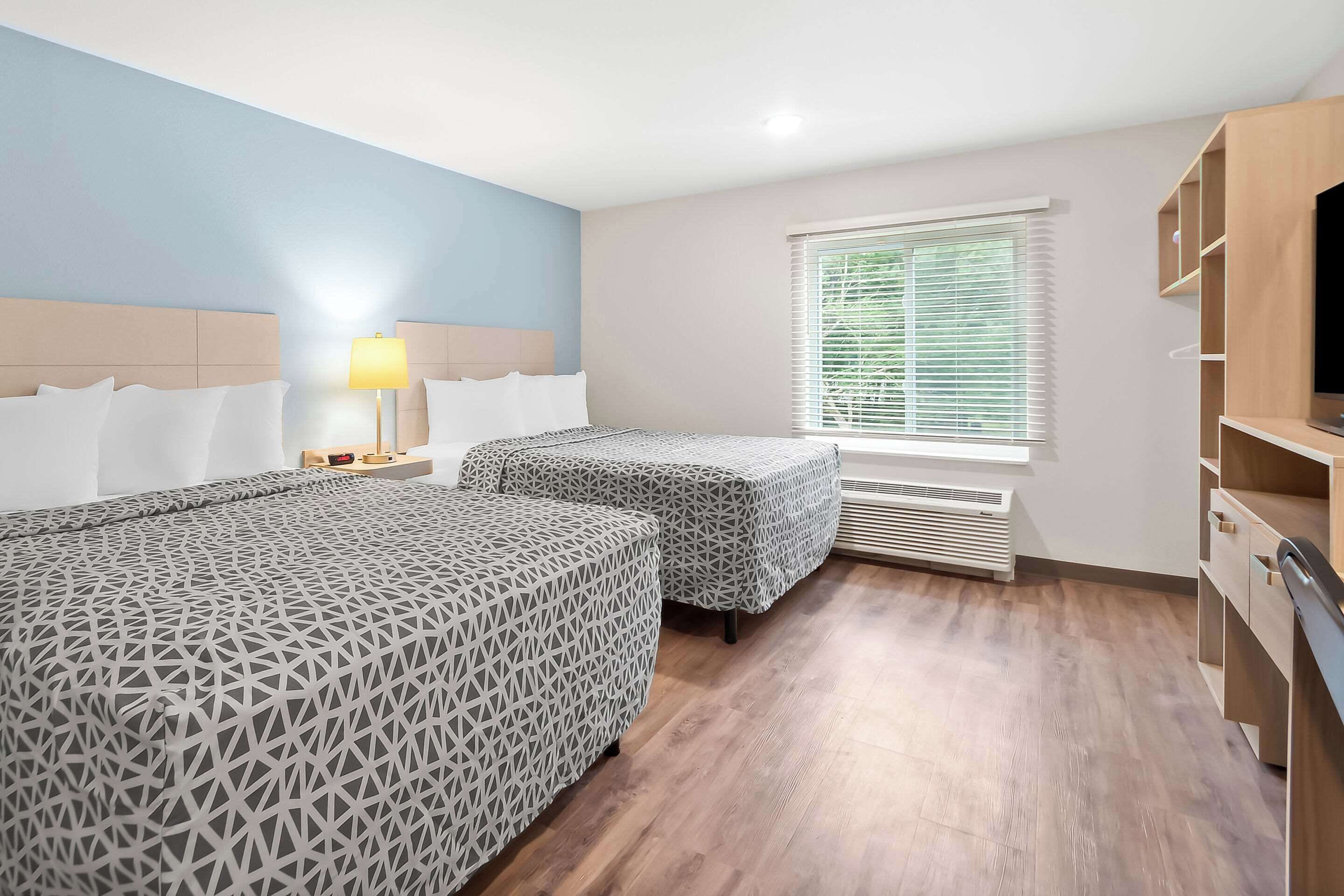 A hotel room with two beds, a nightstand, a lamp, a window with blinds, and a built-in TV shelf.
