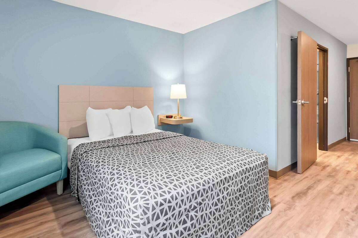 A hotel room with a bed, a nightstand with a lamp, a teal armchair, and a wooden door.