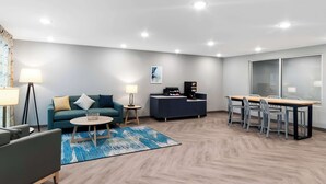 A modern hotel room with a sofa, a small round table, a coffee machine, a desk with chairs, and a large window. - Woodspring Suites Pensacola West (Pensacola)