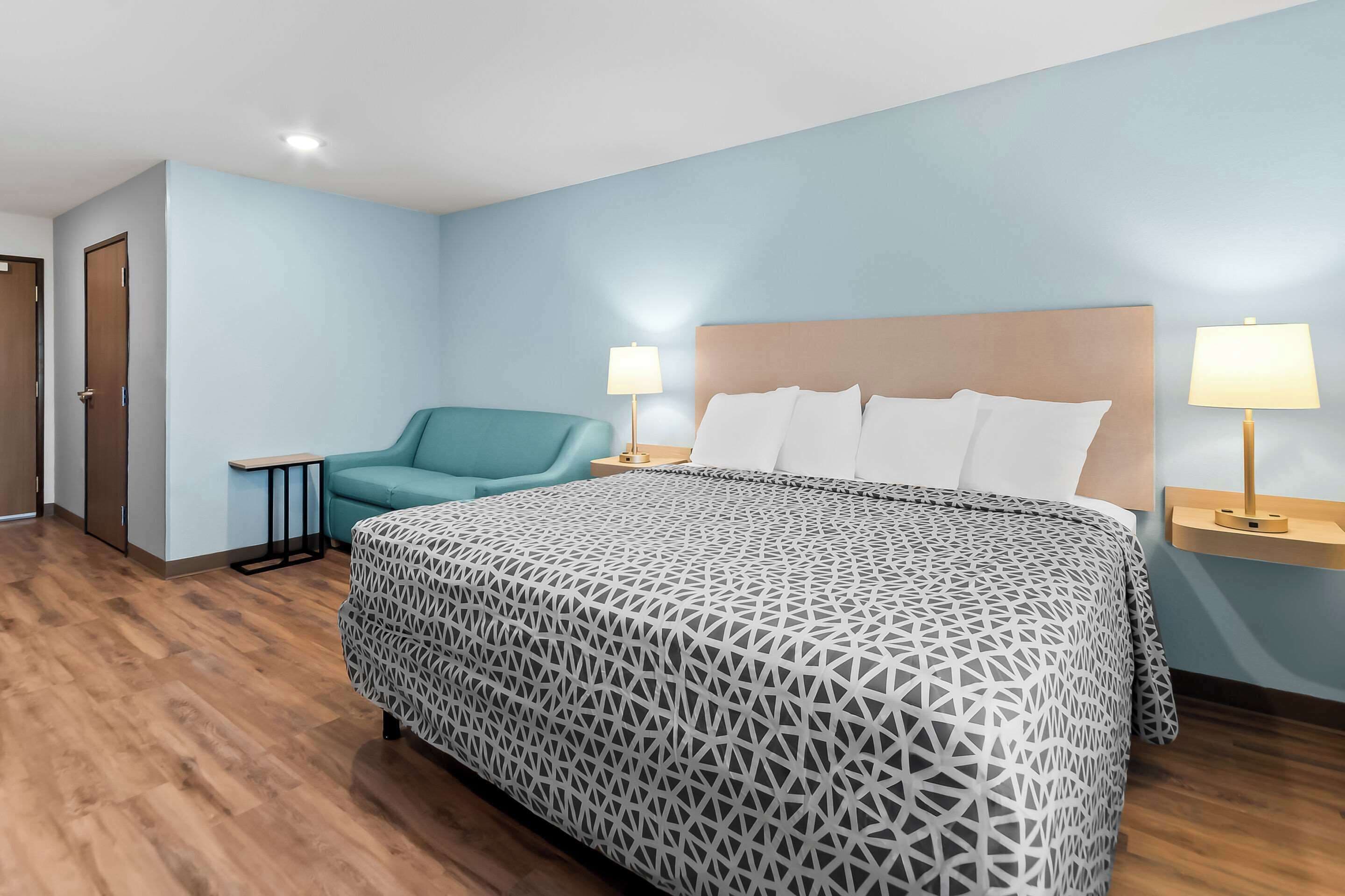 Una habitación de hotel con una cama, una silla color turquesa, dos mesitas de noche con lámparas y suelo de madera.