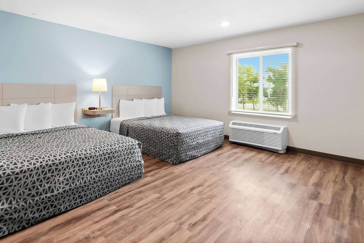 A hotel room with two beds, a nightstand with a lamp, a window with blinds, and an air conditioning unit.