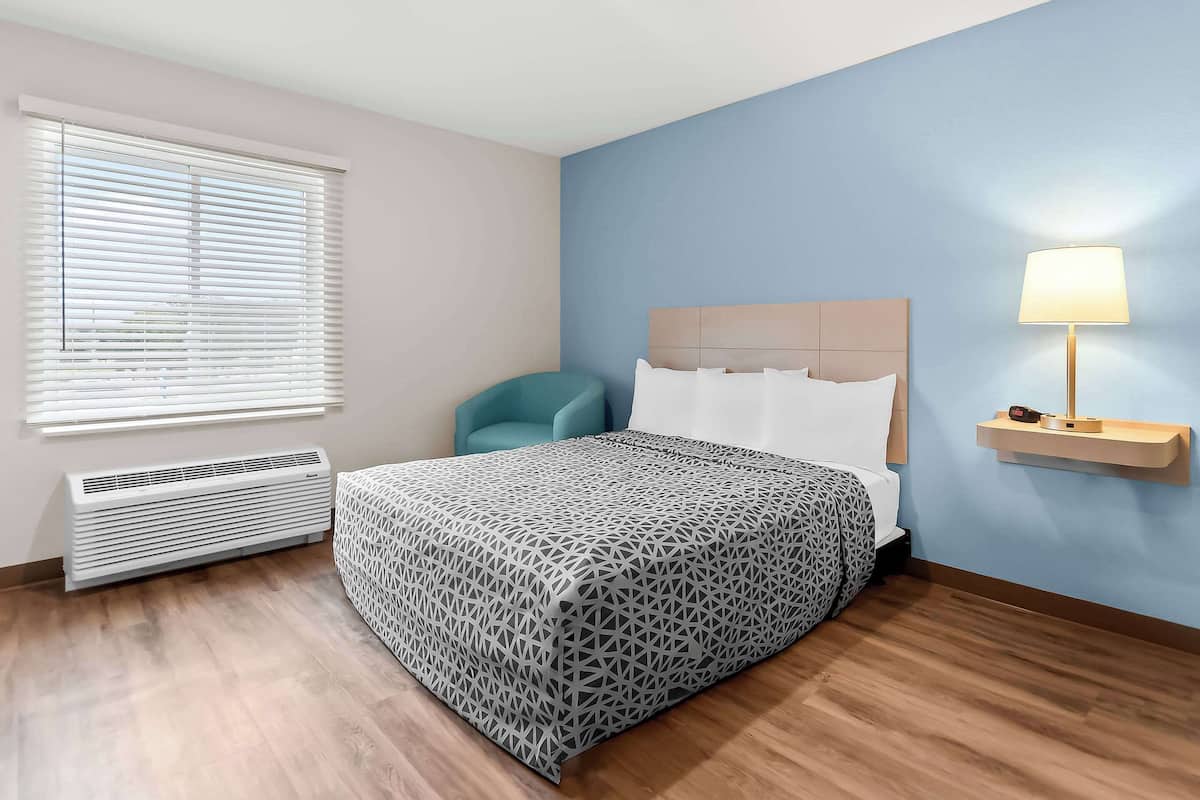A hotel room with a bed, a nightstand with a lamp, a chair, and a window with blinds.