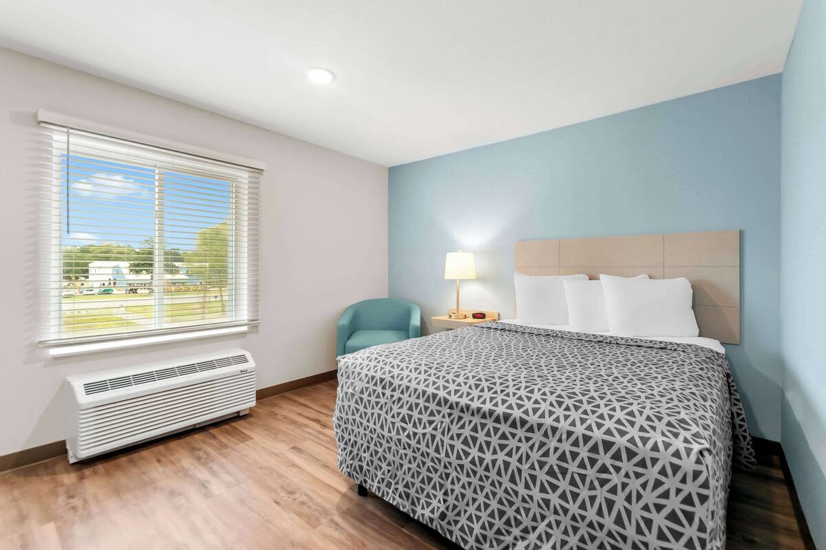 A hotel room with a bed, a chair, a nightstand with a lamp, a window with blinds, and a wall-mounted heater.