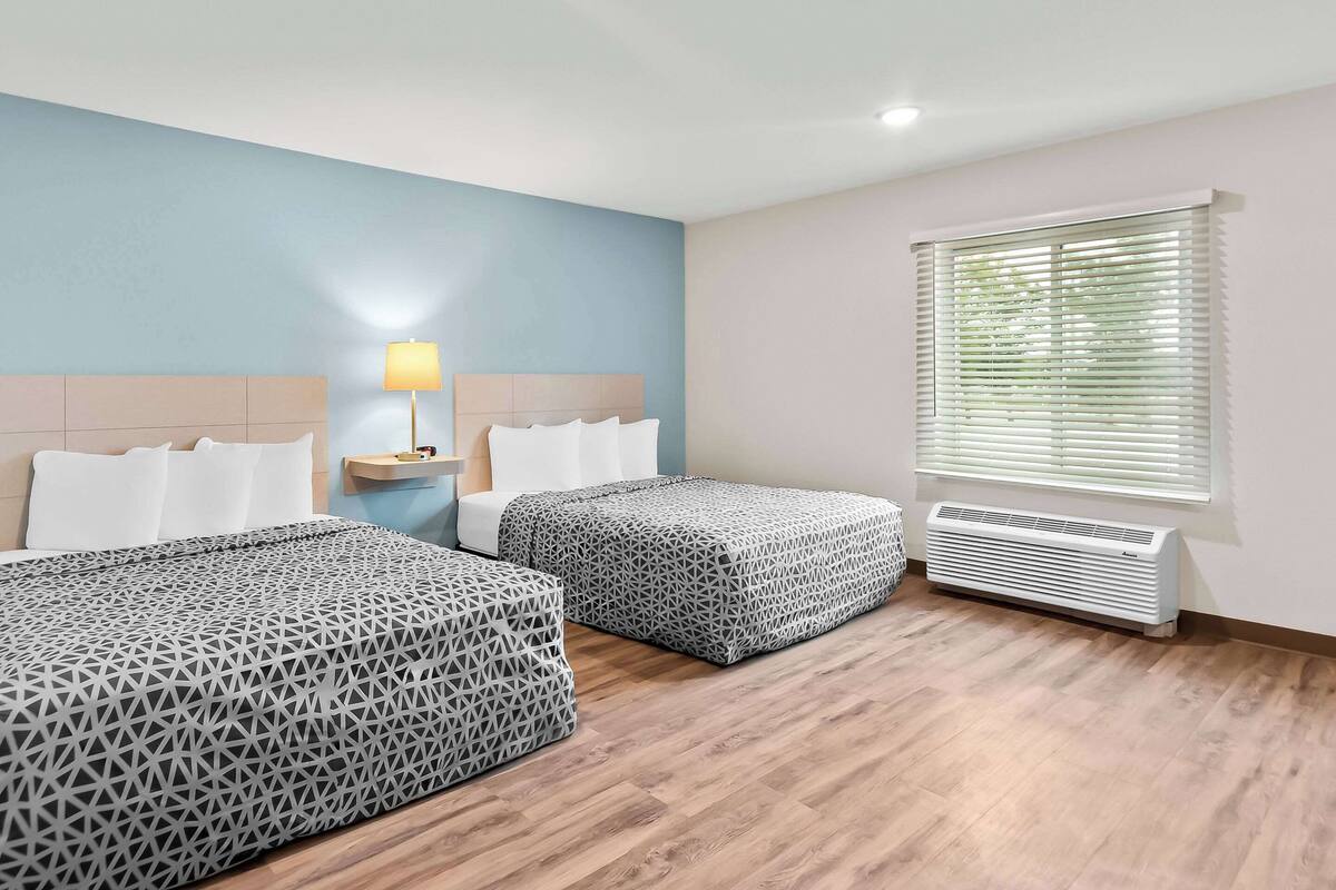 A hotel room with two beds, a bedside table with a lamp, a window with blinds, and a wall-mounted air conditioner.