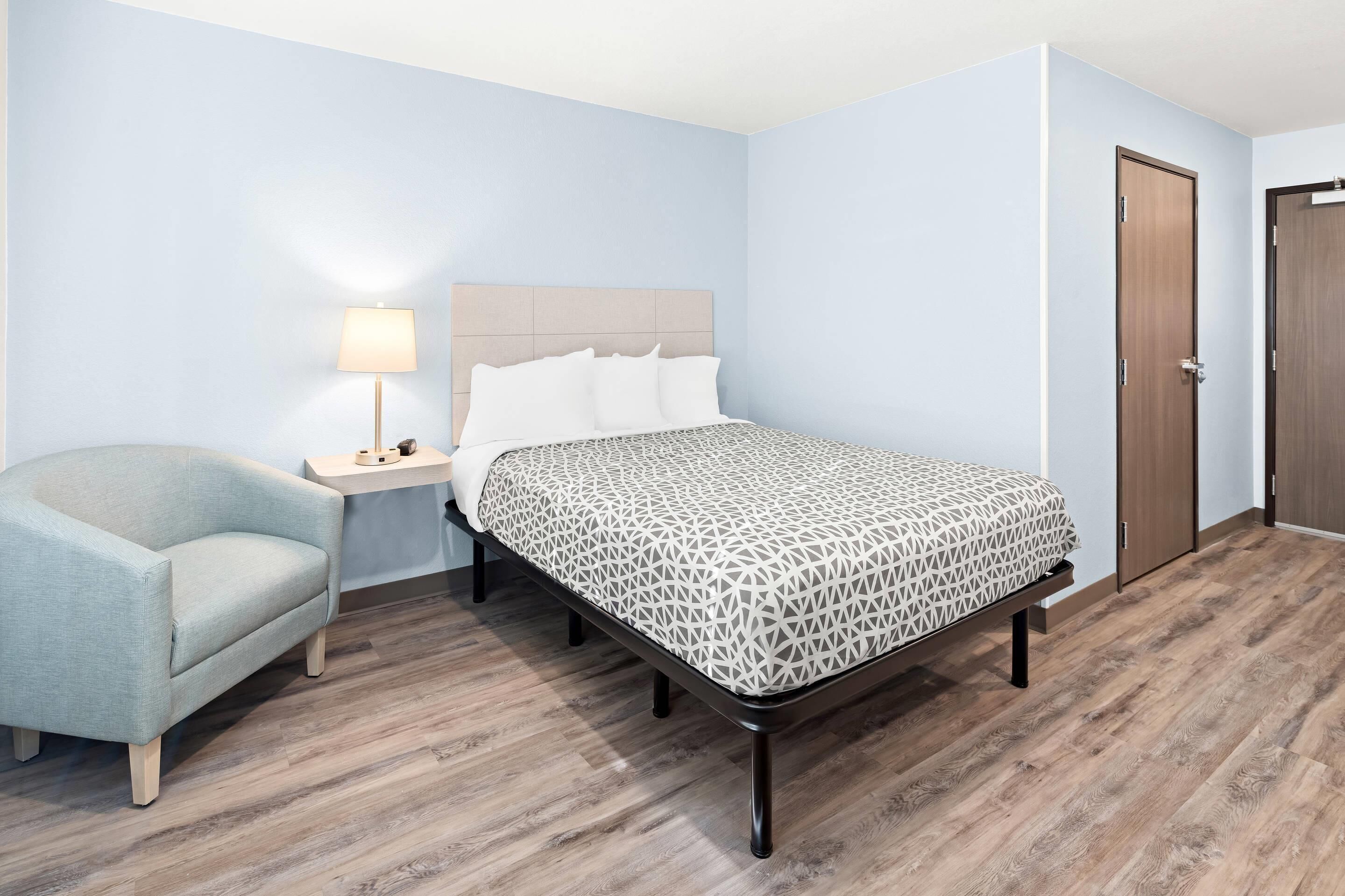 Una habitación de hotel con cama, mesita de noche, lámpara, silla y piso de madera.