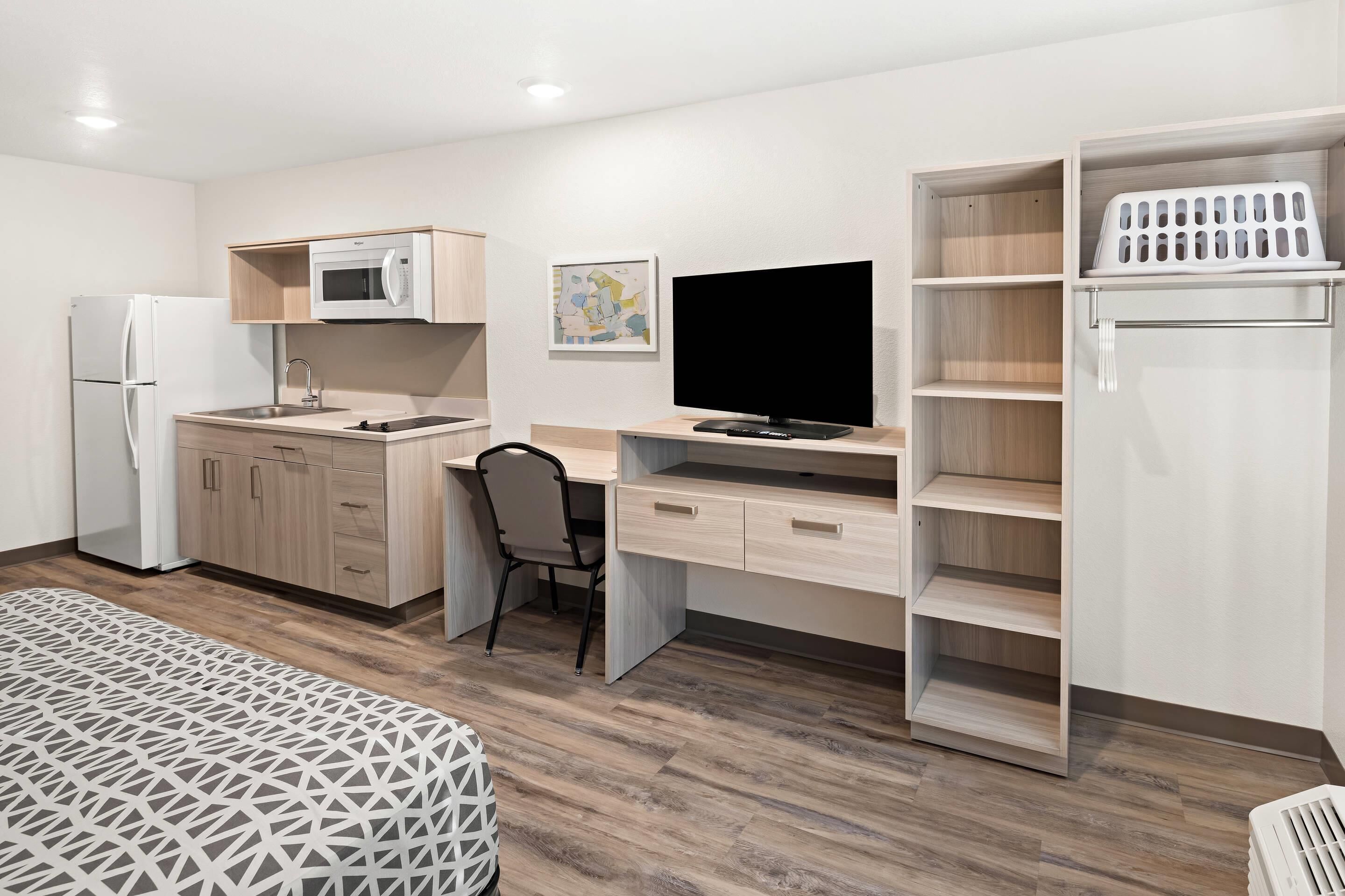 A compact room with a kitchenette, a bed, a desk with a chair, and a TV.