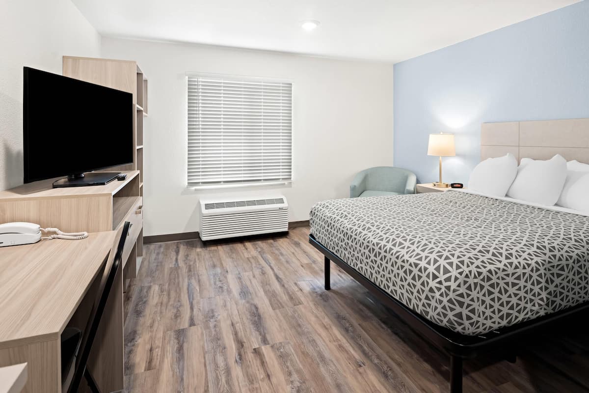 Une chambre d’hôtel comprenant un lit, un bureau avec un téléviseur à écran plat, une chaise, une table de chevet avec une lampe et une fenêtre avec des stores.
