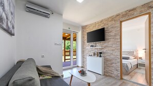 Een moderne woonkamer met een grijze bank, een kleine witte tafel en een televisie die aan een stenen muur bevestigd is. Er is een deur naar een balkon met een houten terras.
