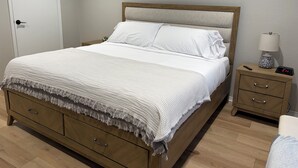 A neatly made bed with white linens and a textured blanket, a wooden nightstand with a lamp and clock, and a door to the left.