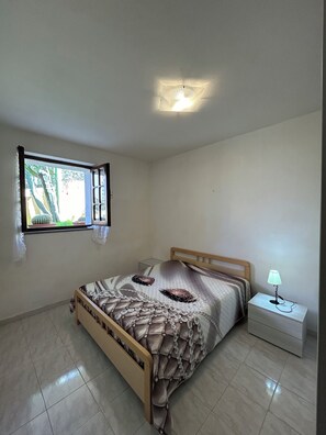 A bedroom with a bed, a nightstand, a lamp, and a window with a view of greenery.