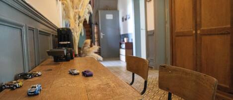 A wooden dining table with toy cars, a chair, and a vase of dried plants in a hallway with patterned flooring and a wooden door.
