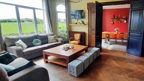 A living room with a sofa, coffee table, and ottomans, a dining area with a table and chairs, and a view of a green landscape through large windows. - Villa BeauSejour in the heart of the countryside  (Baives)