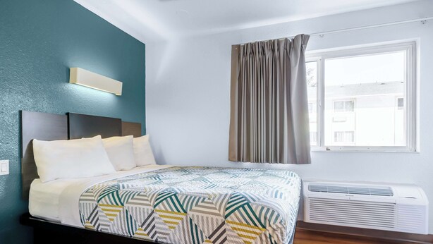 A bedroom with a bed, a wall-mounted air conditioner, a window with curtains, and a view of buildings outside. - Studio 6 Seattle, WA Sea-tac Airport South (SeaTac)