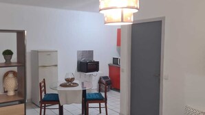 Een functionele keuken met een magnetron, een kleine koelkast en een rode kast. Een eettafel met twee stoelen en een kleine potplant staan links.