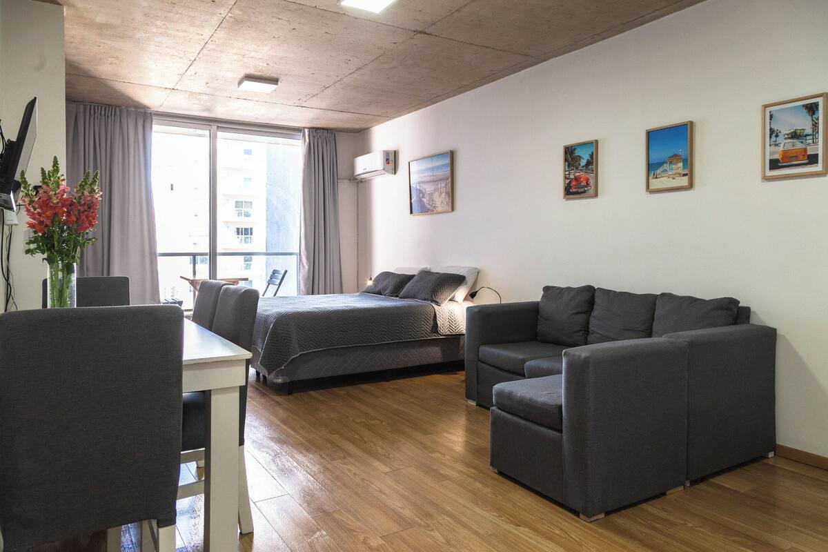 Une chambre d’hôtel moderne avec un lit, un canapé, une table à manger et des chaises. Trois tableaux encadrés ornent le mur, et une fenêtre est agrémentée de rideaux.