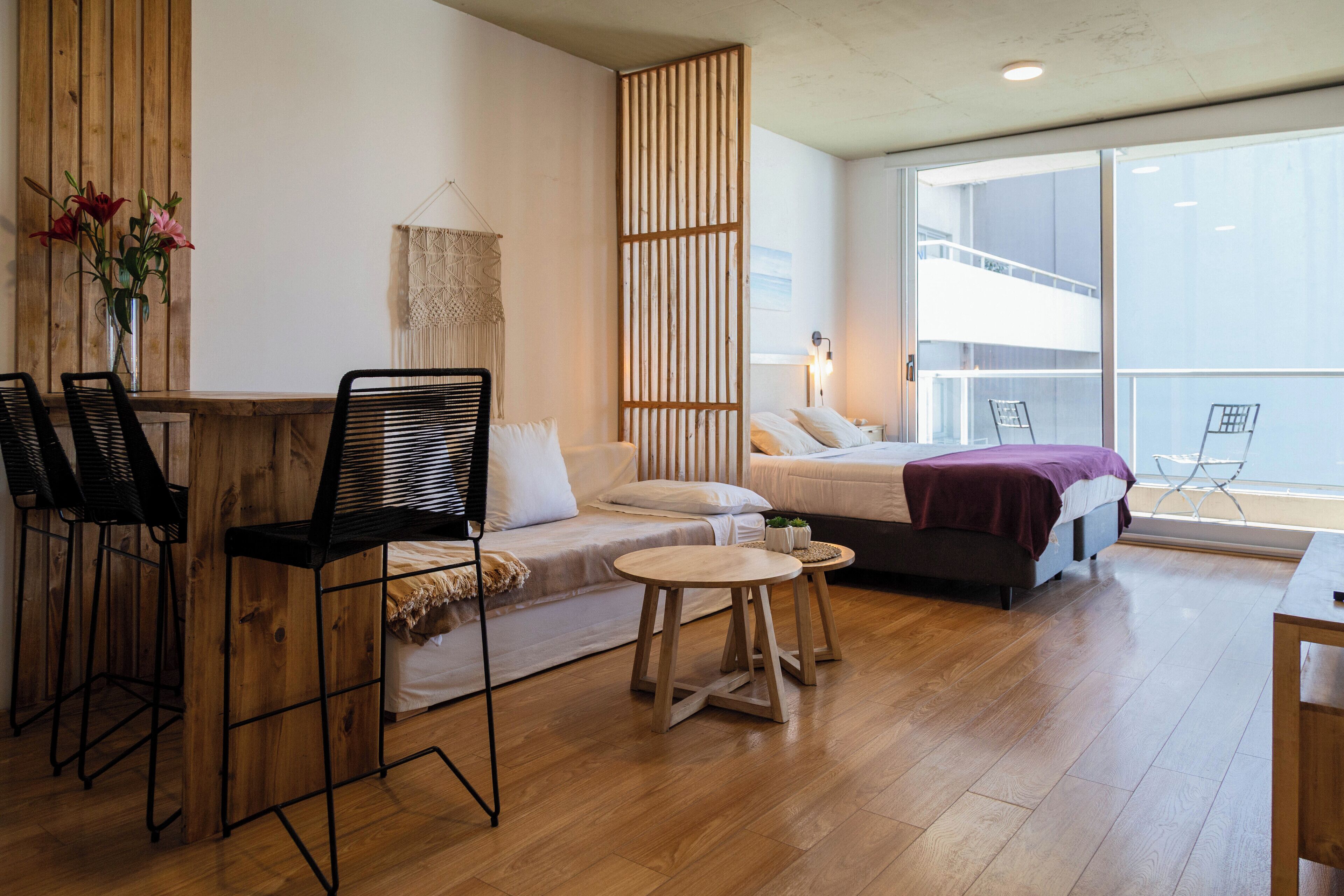 Una habitación moderna con piso de madera, una cama con cabecera blanca, una mesita redonda y dos sillas negras.
