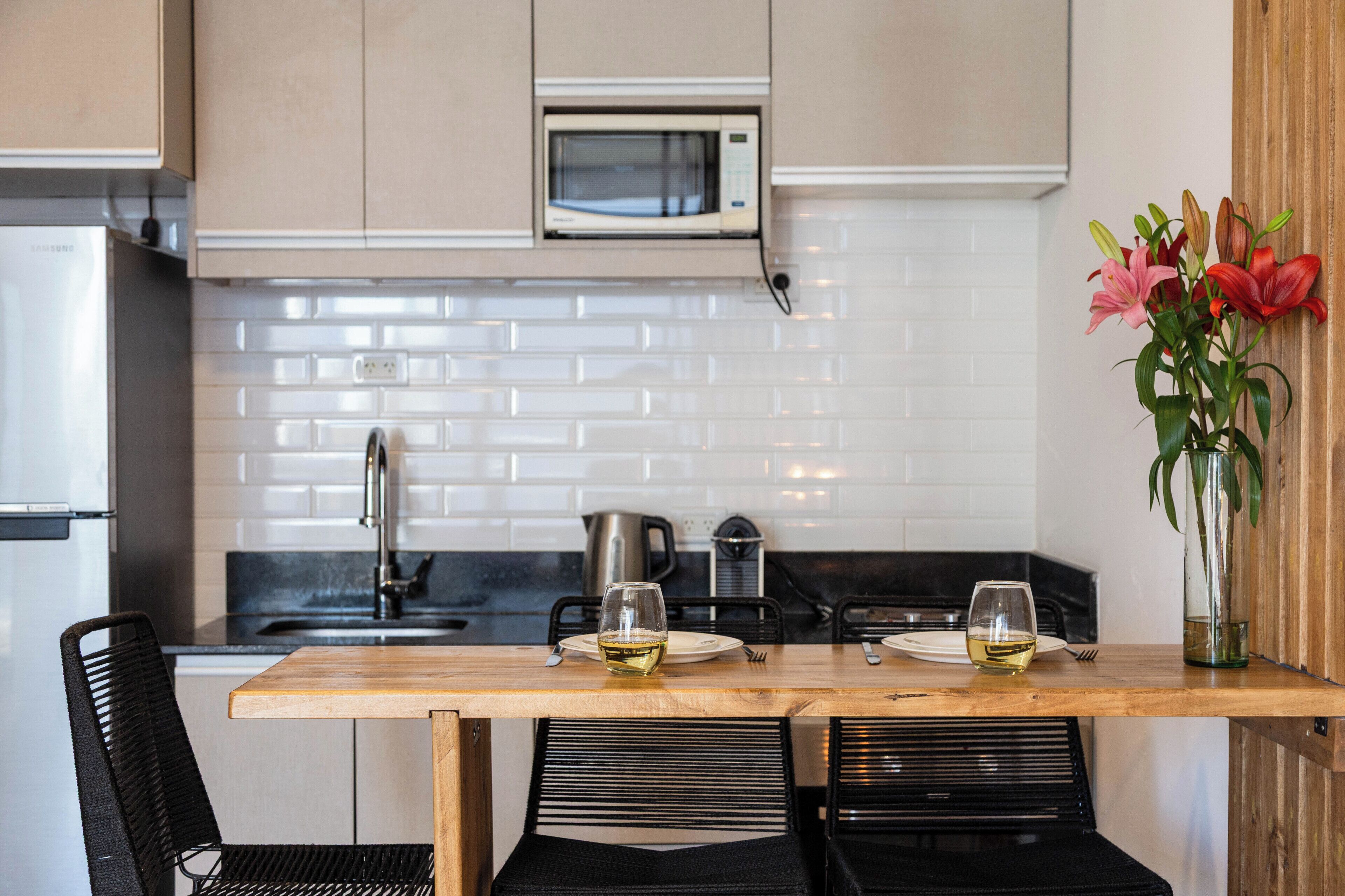 A modern kitchen with a wooden table, two wine glasses, a microwave, and a vase with flowers.