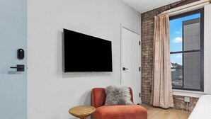 Une chambre moderne avec un téléviseur à écran plat fixé au mur, un fauteuil rouge agrémenté d’un coussin moelleux, une petite table d’appoint en bois et une grande fenêtre avec des rideaux.