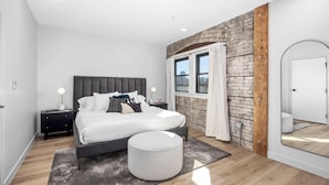 Une chambre à coucher moderne avec un grand lit, un pouf rond, une table de chevet et une fenêtre avec des rideaux.
