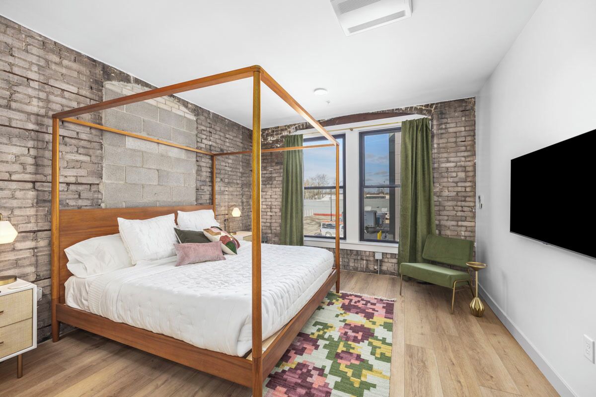 Un dormitorio moderno con cama con dosel, pared de ladrillo, ventana grande y una silla verde.