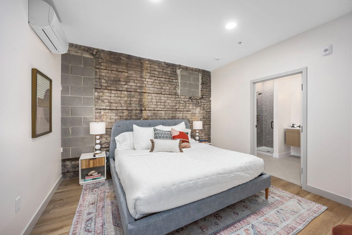 A bedroom with a brick accent wall, a bed with white bedding, a nightstand with a lamp, and a door leading to a bathroom.