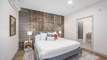 A bedroom with a brick accent wall, a bed with white bedding, a nightstand with a lamp, and a door leading to a bathroom.