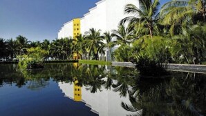 A modern hotel with a white facade and a yellow accent building, surrounded by palm trees and a reflective water body.
