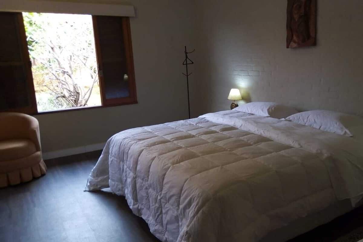 Quarto com cama grande, duas luminárias de cabeceira, uma cadeira e janela com vista para árvores.