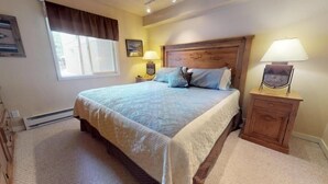 Ein Schlafzimmer mit einem großen Bett, einem Holz-Kopfteil, zwei Nachttischlampen, einem Nachttisch und einem Fenster mit Vorhängen.
