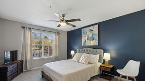 Ein Schlafzimmer mit einem Bett, einem Deckenventilator, einem Fernseher, einem Fenster mit Vorhängen, einem Nachttisch mit Lampe und einem Sessel.