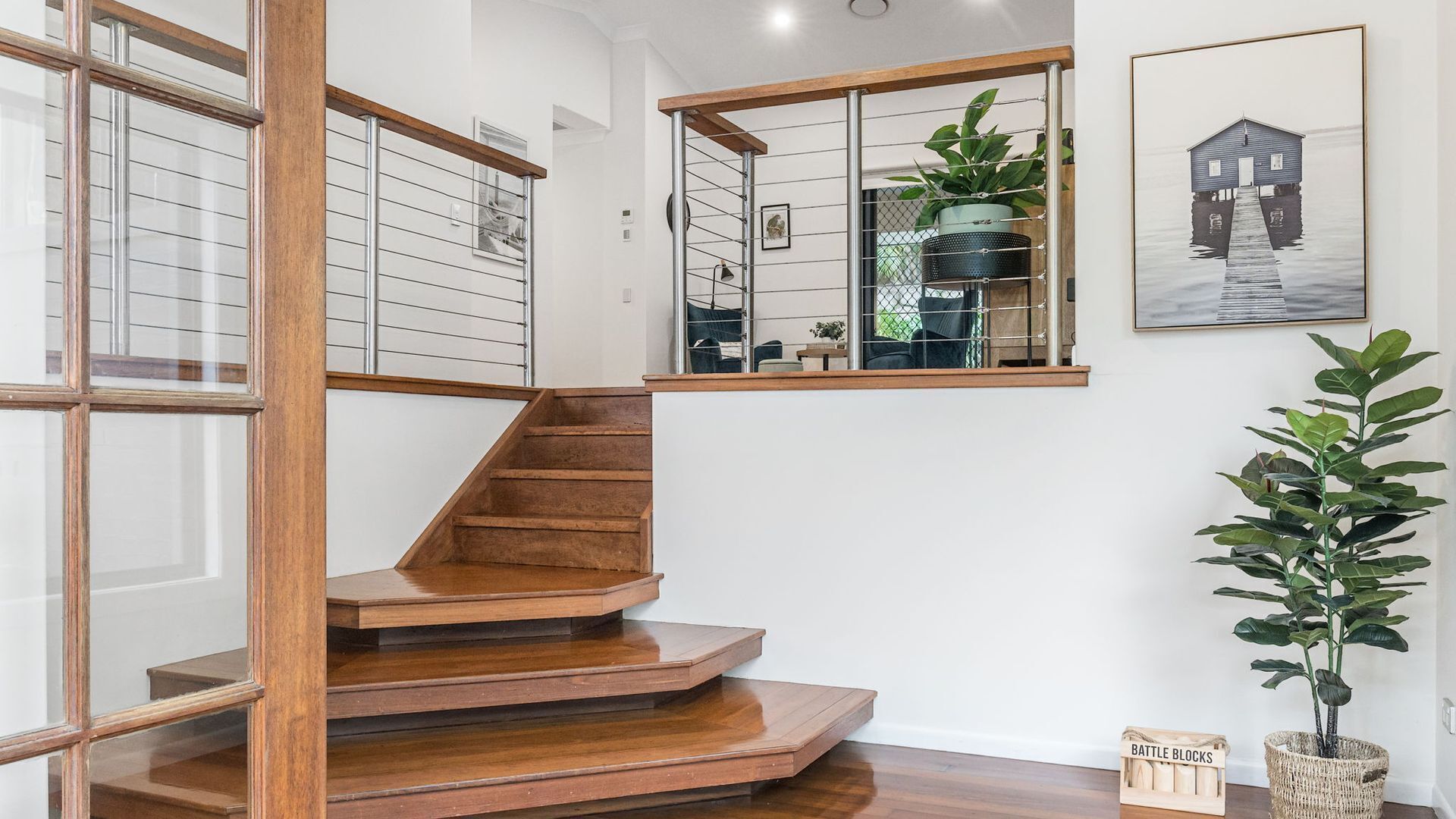 A wooden staircase leading to a second-floor landing with a glass railing, a potted plant, and a framed artwork on the wall.