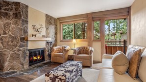 Una sala de estar acogedora con una pared de piedra, chimenea, sillones de cuero, sofá y un ventanal con vistas a árboles.