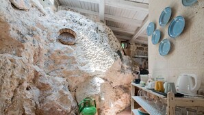 Tunnelmallinen, maalaishenkinen sisustus, jossa on kiviseinät, punottu kori, vihreä keramiikkakannu ja hylly, jossa on lautasia ja keittiötarvikkeita.