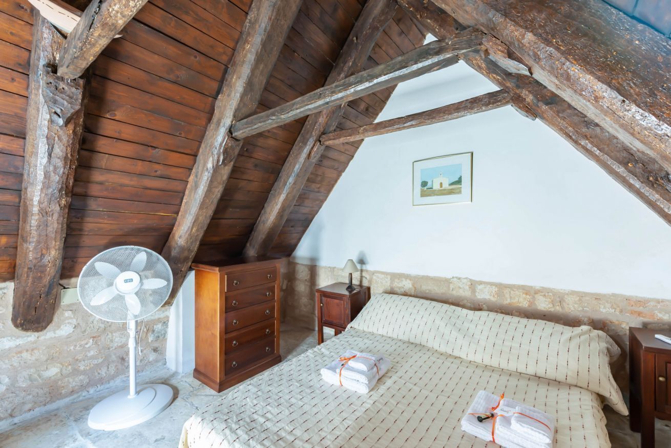 Een slaapkamer met een houten plafond, een bed met een bedsprei met een patroon, een dressoir, een nachtkastje en een aan de muur hangend schilderij.