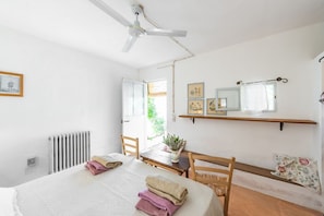 A bedroom with a bed, a ceiling fan, a radiator, a window, a table with chairs, and a shelf with pictures.