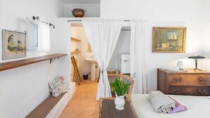 A room with a bed, a bedside table with a lamp, a chair, a small table with a plant, a shelf with a picture, and a door leading to a bathroom.