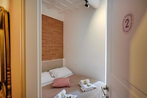 A bedroom with a brick wall, a bed with white linens, a pink pillow, and towels on the side.