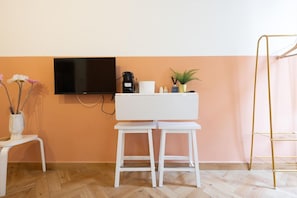 A room with a TV mounted on the wall, a white desk with two stools, a coffee maker, a small plant, and a gold ladder against the wall.