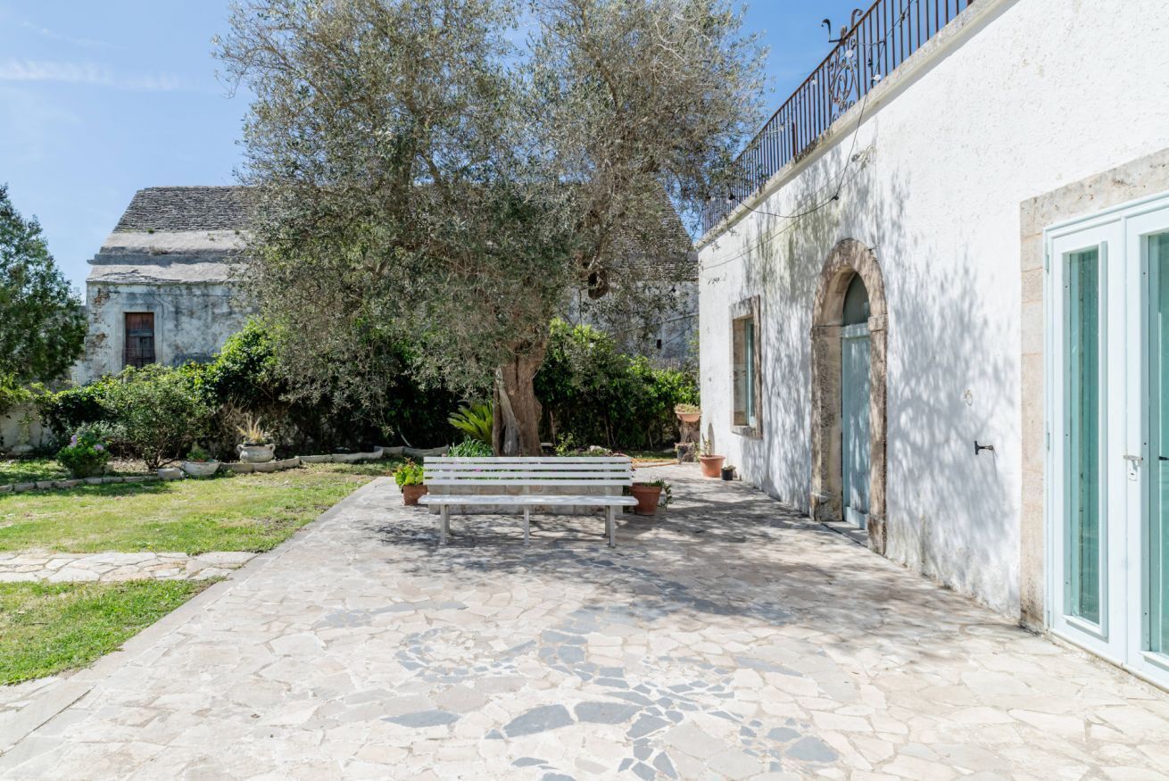 A courtyard with a stone bench, a white building with an arched doorway, and a tree.