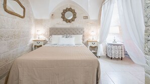 A bedroom with a large bed, a decorative mirror, a round mirror, a nightstand, and a small table with a lace tablecloth. - Portici Guest House by Rentbeat (Fasano)