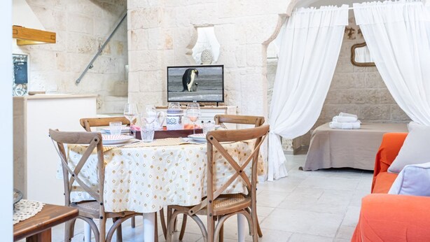 A dining area with a table set for two, a television displaying an image of a penguin, and a cozy seating area with a bed and a towel. - Portici Guest House by Rentbeat (Fasano)