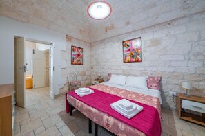 A bedroom with a stone wall, a bed with a pink and white bedspread, two paintings, a nightstand, and a wooden desk.