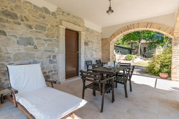 A stone-walled room with a bed, a table with chairs, and an archway leading to an outdoor area with greenery. - Nonna Assunta by Rentbeat (Abbateggio)