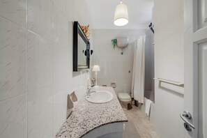 A modern bathroom with a granite countertop sink, a wall-mounted mirror, a toilet, and a shower area. - Piermarie by Rentbeat (Ostuni)