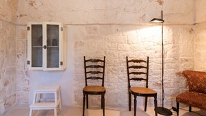 A room with a rustic stone wall, two wooden chairs, a small white stool, a cabinet with glass doors, and a floor lamp. - Masseria Fumarola Il Cavaliere IV by Rentbeat (Ostuni)